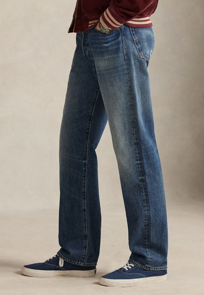 Plave traperice opuštenog kroja, s izblijedjelim efektom i blagim tragovima habanja. Uparene s plavim tenisicama.