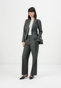 Woman wearing a dark gray pantsuit with a white shirt, black pointed shoes, and a black shoulder bag, standing against a white background.