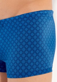 Shorts de bain à motif bleu avec un design en losange dans des teintes de bleu variées, un ajustement snug et une texture de tissu lisse.