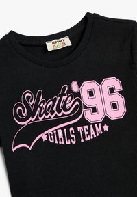 Sort ribbet t-shirt med pink grafisk tekst: "Skate' 96" og "GIRLS TEAM." Rund halsudskæring, blød tekstur og korte ærmer.