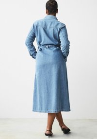 Denim-paitapuku vaaleansinisellä värillä, pitkillä hihoilla, vyötäröllä vyö ja polven keskivaiheilla. Varustettu kauluksella ja metallinapit yksityiskohtina.