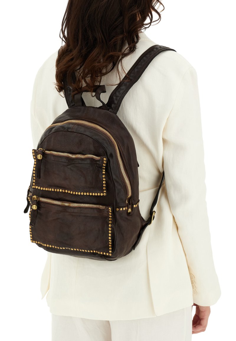 Zaino in cuoio marrone con accenti in oro a forma di borchie, tasche con doppia zip sul davanti e spallacci imbottiti, indossato sopra un blazer bianco.
