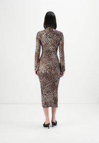 Woman with straight black hair wearing a long-sleeve, leopard print midi dress and black heeled mules, standing against a plain white background.