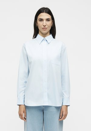 Žena s rovnými tmavými vlasmi, oblečená v svetlomodrej košeli na gombíky a svetlomodrých rifliach, stojaca na jednoduchom bielom pozadí.