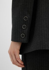 Manica di un blazer grigio scuro a righe sottili con tre bottoni circolari in argento, texture liscia e sartoria accurata. Mano parzialmente visibile.