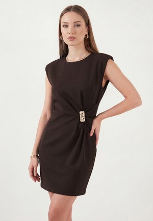 Kvinde iført en sort ærmeløs kjole med en guldbælte-detalje i taljen, poserer med den ene hånd på hoften mod en ensfarvet baggrund.