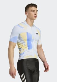 Radsporttrikot in Hellblau mit gelben und blauen geometrischen Mustern, kurzen Ärmeln und einem Reißverschluss vorne. Kombiniert mit schwarzen, enganliegenden Shorts.