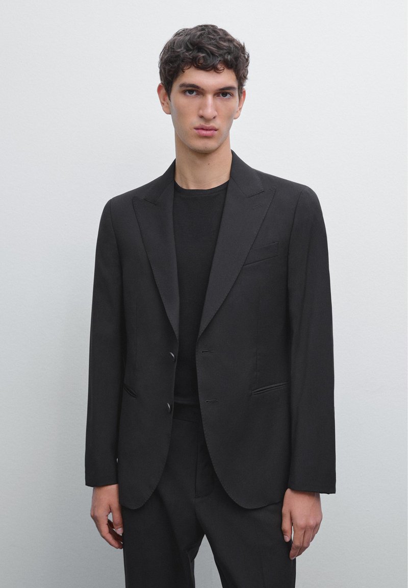 Schwarze, taillierte Blazerjacke mit Reverskragen, zwei Knöpfen und seitlichen Taschen. Glattes Material und strukturierter Schnitt. Über einem schwarzen T-Shirt getragen.