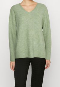 Pull en maille côtelée vert clair avec col en V et manches longues, présentant une coupe décontractée et une texture douce, associé à un pantalon noir texturé.