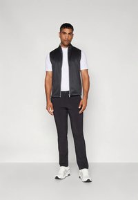 Schwarze Weste mit Reißverschluss über einem weißen Polohemd, kombiniert mit schwarzen Hosen und weißen Turnschuhen. Die Weste zeichnet sich durch ein elegantes, glattes Design aus.