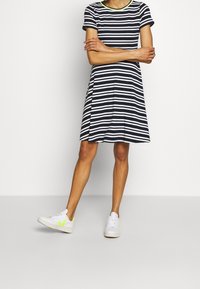 Robe T-shirt rayée bleu marine et blanche avec des manches courtes, une jupe évasée et un col vert. Associée à des baskets blanches avec des accents néon.