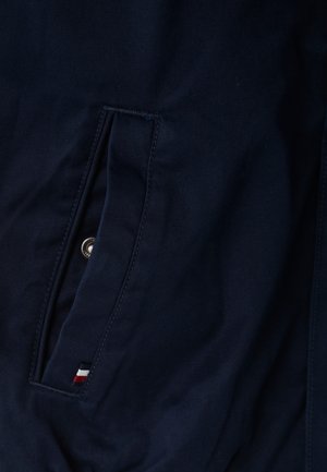 Chaqueta de tela azul marino con una textura diagonal prominente, con un bolsillo lateral y un pequeño logotipo en rojo, blanco y azul.
