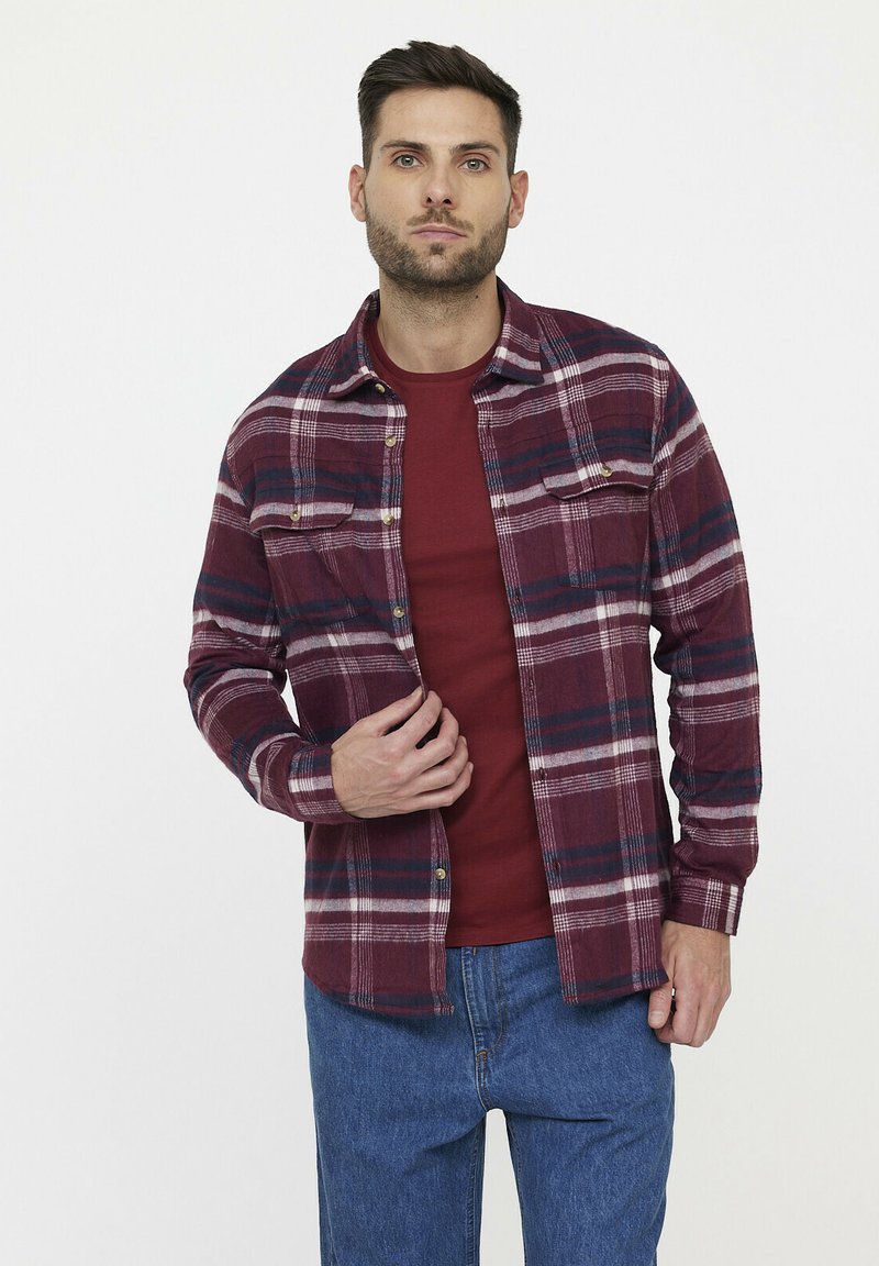 Chemise en flanelle à carreaux rouge et marine, avec un devant à boutons, deux poches poitrine et des manches longues, superposée à un t-shirt à col rond rouge.