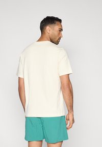 Κοντό μπεζ μπλουζάκι με κοντά μανίκια και στρογγυλή λαιμόκοψη, σε συνδυασμό με τιρκουάζ σορτς, από πίσω όψη, με απλό σχέδιο.