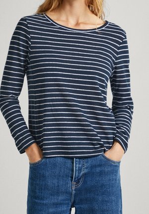 Vrouw draagt een marineblauw-wit horizontaal gestreept shirt met lange mouwen, lichtjes ingestopt in een hooggetailleerde blauwe spijkerbroek, handen in de zakken.