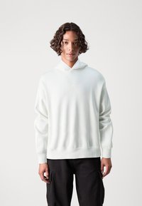 Λευκή φούτερ με κουκούλα, χαλαρή εφαρμογή, ριμπ μανσέτες και στρίφωμα. Μαλακή υφή, μινιμαλιστικός σχεδιασμός, συνδυασμένη με μαύρα cargo παντελόνια.