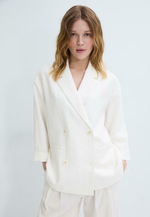 Jeune femme aux cheveux châtain clair portant un blazer blanc à double boutonnage ample et un pantalon assorti sur un fond uni.