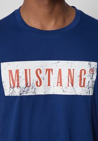 Blaues T-Shirt aus Baumwolle mit einem großen weißen rechteckigen Grafikdesign mit strukturierten Kanten und dem Wort "MUSTANG" in fetten orangen Buchstaben.