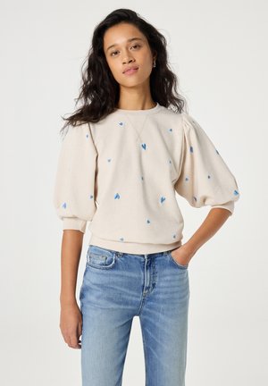 Vrouw met lang donker haar draagt een beige sweatshirt met pofmouwen en blauwe hartjespatronen, gecombineerd met lichtblauwe spijkerbroek, staat met één hand in de zak.