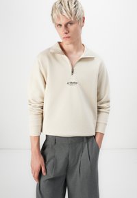 Kerman fleece-pullover dengan simbol hitam di dada, dilengkapi dengan ritsleting seperempat. Celana abu-abu plis menjadi pelengkap penampilan.