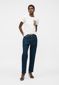 Γυναίκα στέκεται φορώντας λευκό t-shirt Moschino με εκτύπωση αρκουδάκι, σκούρο μπλε τζιν, μαύρη ζώνη και μαύρα σανδάλια με ψηλό τακούνι μπροστά σε λευκό φόντο.