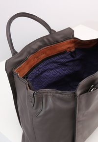 Grå läderhandväska med dragkedjestängning, brunt mockaskärp och ett lila inrede med vågmönster. Dubbla handtag för bärande.