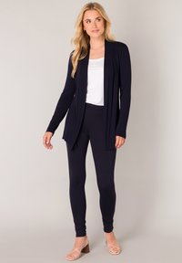 Marineblauwe lange mouw cardigan over een witte top, gecombineerd met marineblauwe legging en lichtbruine sandalen met bandjes. Soepele, rekbare stof met een ontspannen pasvorm.