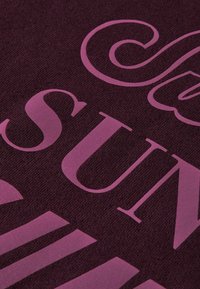 Textil en burdeos oscuro con grandes letras impresas en rosa y acabado suave y mate. El texto incluye elementos estilizados y diferentes tamaños de fuente.