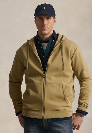 Mand iført tan lynlås hættetrøje over grønternet skjorte, blå jeans, navy baseballkasket og armbåndsur, stående mod neutral baggrund.