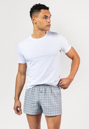 Graue Boxer mit schwarzem Karomuster, ausgestattet mit einem elastischen Bund und einem Knopf vorne. Kombiniert mit einem figurbetonten weißen T-Shirt.