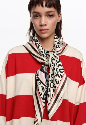 Mujer joven con cabello corto y oscuro que lleva un top de rayas rojas y blancas y una bufanda de color claro con un patrón abstracto en negro y rosa atada alrededor del cuello.