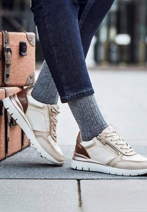 Persona con zapatillas beige y marrones, calcetines grises acanalados y vaqueros azul oscuro, de pie cerca de una maleta marrón vintage sobre la acera.