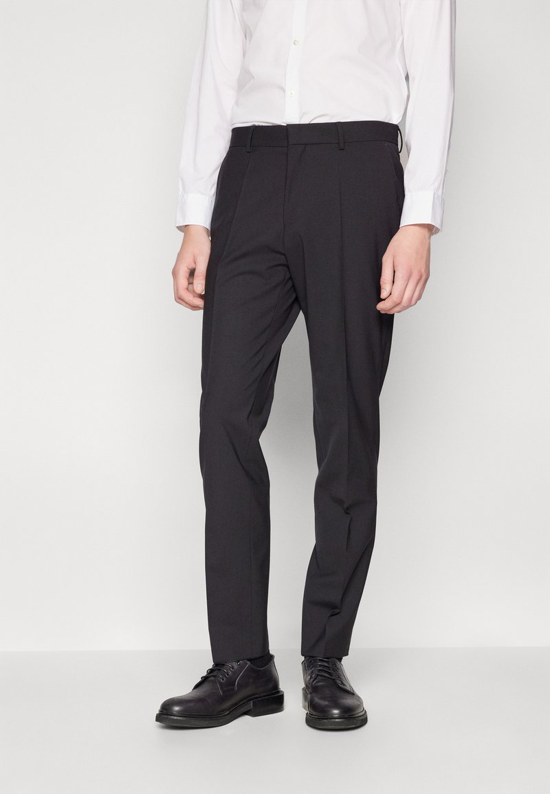 Schwarze, taillierte Hose mit schlanker Passform, ausgestattet mit einer glatten Textur, Gürtelschlaufen und einer Bügelfalte auf der Vorderseite, kombiniert mit schwarzen Schuhen.