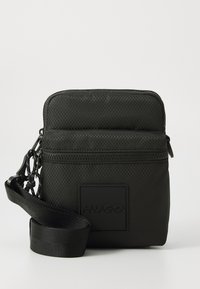 Black textured nylon bag with zip closure, featuring a front logo patch, adjustable strap, and a chain accent attached to the zipper.