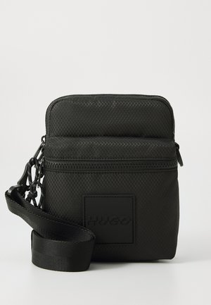 Black textured nylon bag with zip closure, featuring a front logo patch, adjustable strap, and a chain accent attached to the zipper.