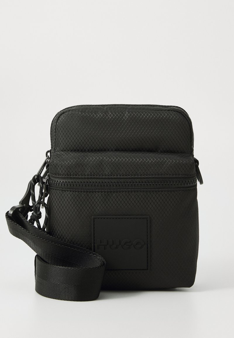 Black textured nylon bag with zip closure, featuring a front logo patch, adjustable strap, and a chain accent attached to the zipper.