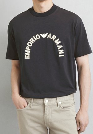 Czarna bawełniana koszulka z dużym, łukowatym, białym logo "EMPORIO ARMANI" na przodzie. Ma okrągły dekolt i luźny krój, zestawiona z beżowymi dżinsami.