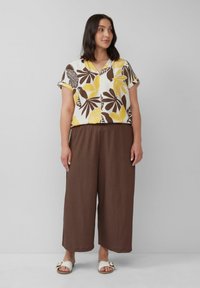 Kurzärmlige V-Ausschnitt-Bluse mit einem gelben und braunen Blattmuster, kombiniert mit braunen, weit geschnittenen Hosen und weißen Sandalen mit einem Schnallenakzent.