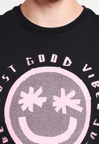 Musta puuvillapaita, jossa on suuri vaaleanpunainen graafinen kuvasto, jossa on hymyilevä kasvot ja tähtisilmät. Teksti kuuluu "Just Good Vibes" paksulla tyylitellyllä fontilla.