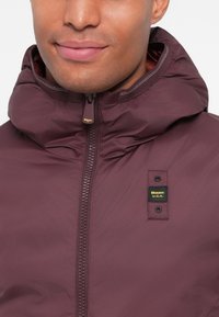 Chaqueta con capucha de color burdeos, con cremallera frontal y una etiqueta de marca. El material parece ligero y la textura es suave con un ligero brillo.