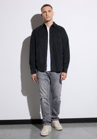 Camisa negra de botones sobre una camiseta blanca, jeans grises y zapatillas beige. La camisa cuenta con un bolsillo en el pecho y un ajuste relajado.