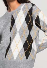 Grauer Pullover mit einem Argyle-Muster aus schwarzen, weißen und gelben Rauten. Das Material wirkt weich und hat eine strukturierte Oberfläche.