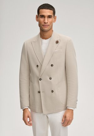 Mężczyzna ubrany w beżową, teksturowaną, dwurzędową marynarkę z sześcioma guzikami, biały sweter i białe spodnie na jednolitym tle.