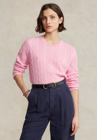 Jeune femme aux cheveux bruns courts portant un pull rose en maille torsadée, pantalon bleu marine taille haute, ceinture noire et bracelets, debout avec une main dans la poche.