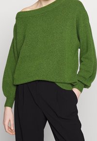 Groene gebreide trui met een off-shoulder ontwerp, lange mouwen en een relaxte pasvorm, gecombineerd met zwarte broeken. Zachte textuur en casual stijl.