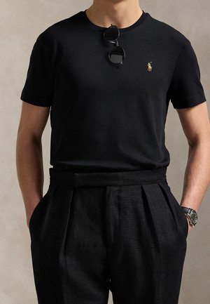 Homme portant un t-shirt noir à manches courtes avec un petit logo brodé, un pantalon noir plissé, des lunettes de soleil rondes suspendues au col et une montre-bracelet.