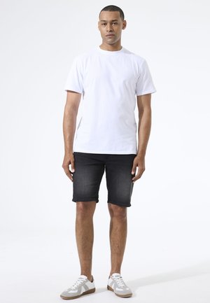 Mann steht vor einem schlichten weißen Hintergrund, trägt ein einfaches weißes T-Shirt, dunkle Jeansshorts und weiße Turnschuhe.