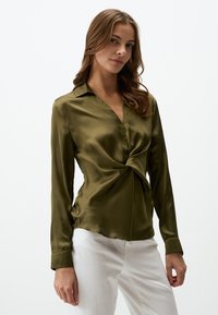 Blouse en satin vert olive avec un détail torsadé à l'avant, un col et des manches longues. Associée à un pantalon blanc, texture lisse et coupe ajustée.