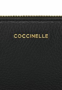 Svart skinnlommebok med teksturert overflate og gull-embossert logo "COCCINELLE." Lukkemekanismen er en metallglidelås med gullakenter.