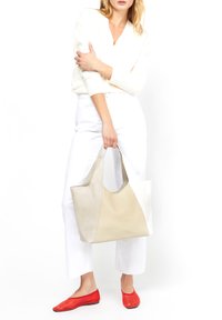 Beige läderhandväska med en slät yta, med kontrasterande paneler och robusta handtag, parat med vita byxor och röda ballerina skor.